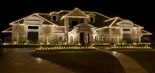 A large stone house at night with warm white lights along every roof line and the driveway edge