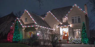 A two-story house at night with red, green and white lights on the roof and two lit trees out front