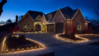 A brick house at dusk with warm white lights along the roof and both sides of the driveway