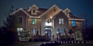 A two-story stone house at night with warm white lights along the roof lines and a wreath over the door