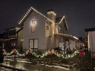 A white farmhouse at night with warm white lights on the roof lines and lit garland along the fence