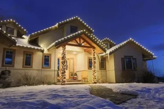 A craftsman house in snow with warm white lights along the roof lines and the porch