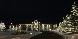A ranch house at night with warm white lights along the roof and lit evergreen trees in the yard