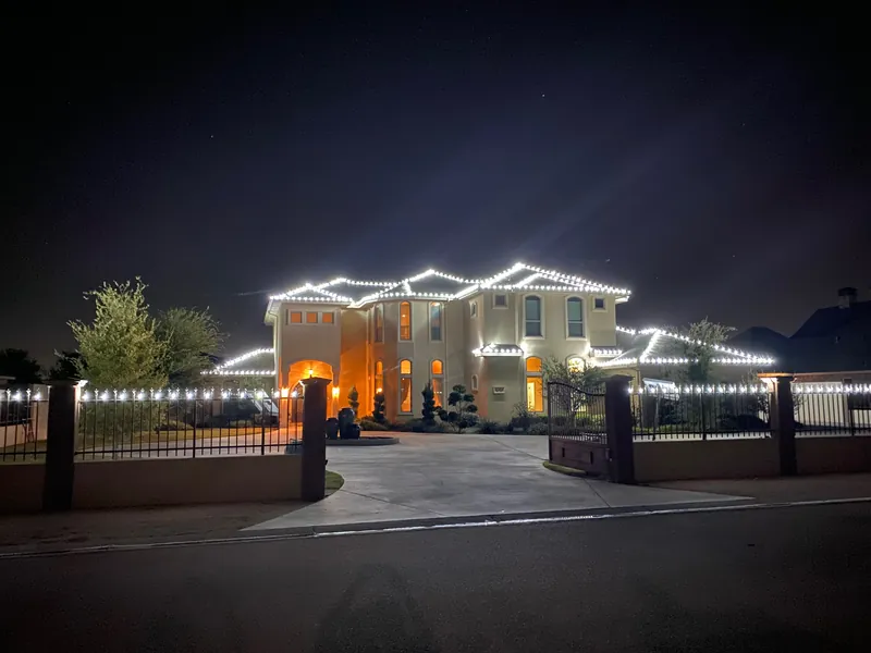 A large two-story house at night with warm white lights along the roof lines, the fence and the gate posts