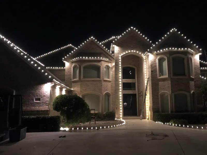A large brick two-story house at night with warm white C9 bulbs along every roof peak, the entry arch and the flower bed